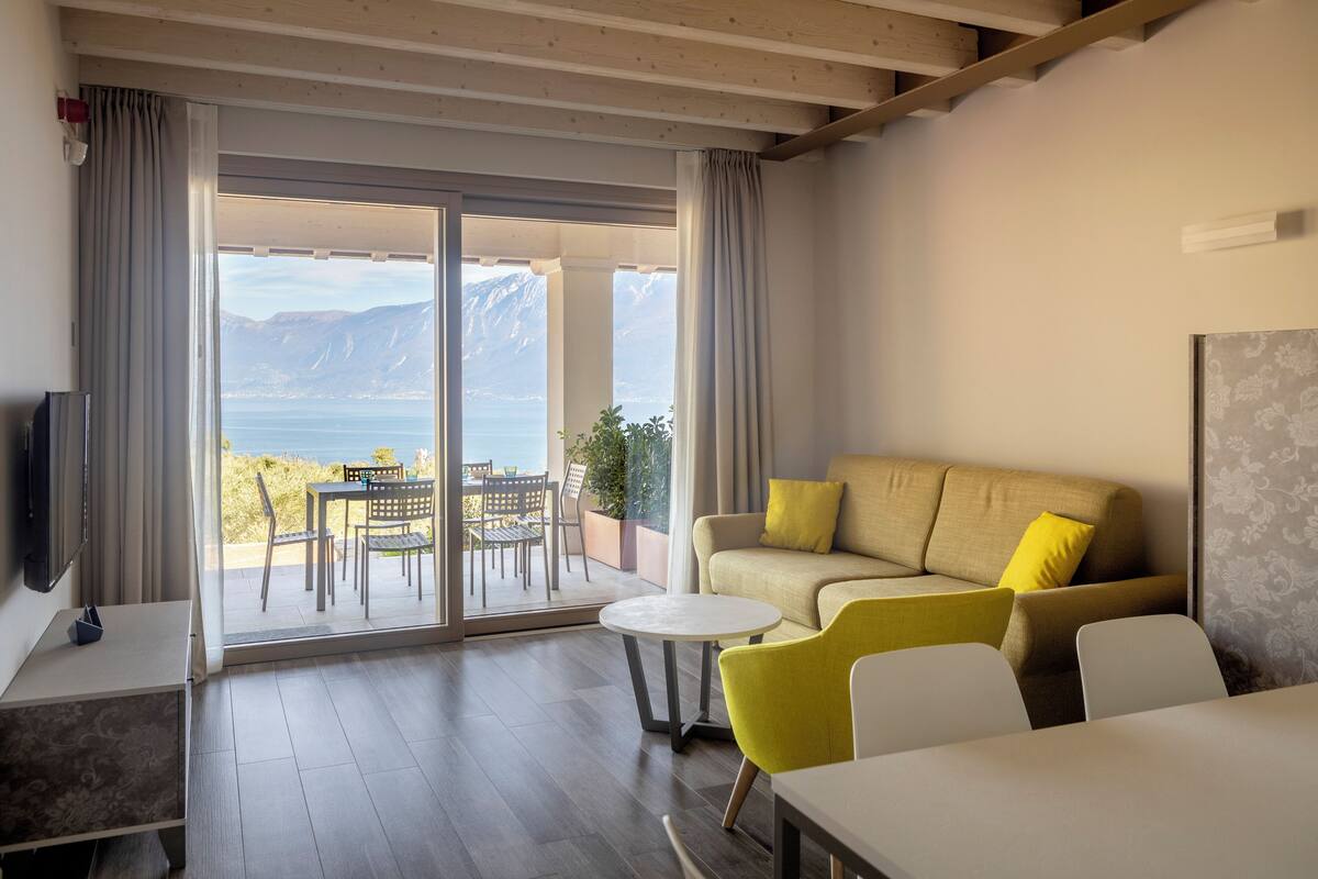 A modern living room with a sofa, chairs, a TV, and a table. It has a large sliding glass door leading to an outdoor area with a view of mountains and a body of water.