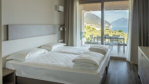 A hotel room with a large bed, a nightstand, a dresser, and a balcony with a view of the sea and mountains. - Relais Zenner (Toscolano Maderno)