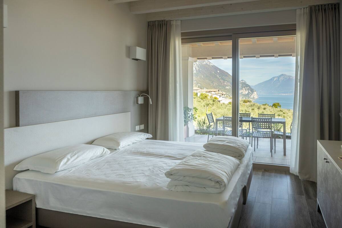 A hotel room with a large bed, a nightstand, a dresser, and a balcony with a view of the sea and mountains.