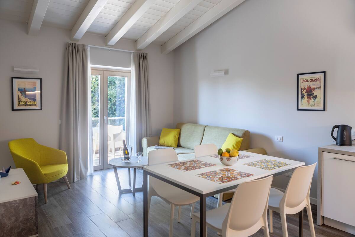 A modern living room with a sofa, a dining table with chairs, a coffee machine, and a window with curtains.