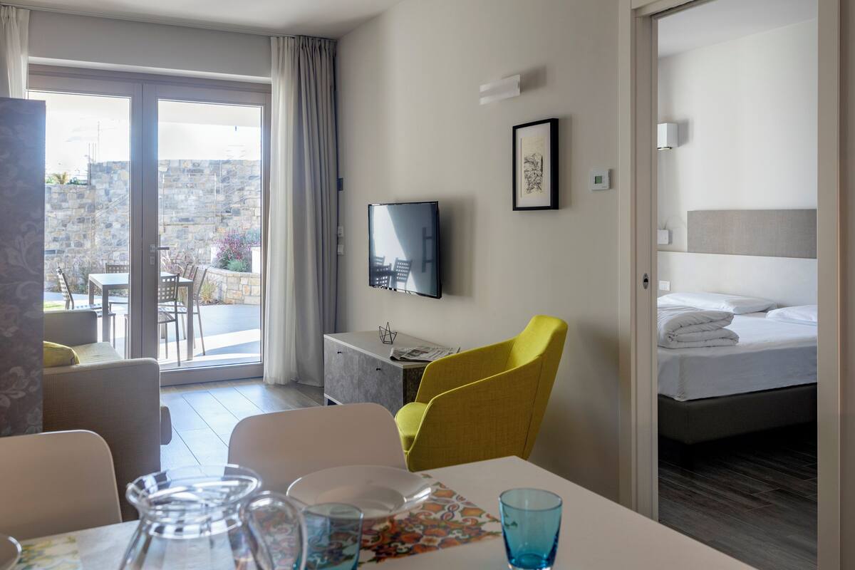 A hotel room with a bed, a desk with a flat-screen TV, a dining table with chairs, and a balcony with a view of the city.