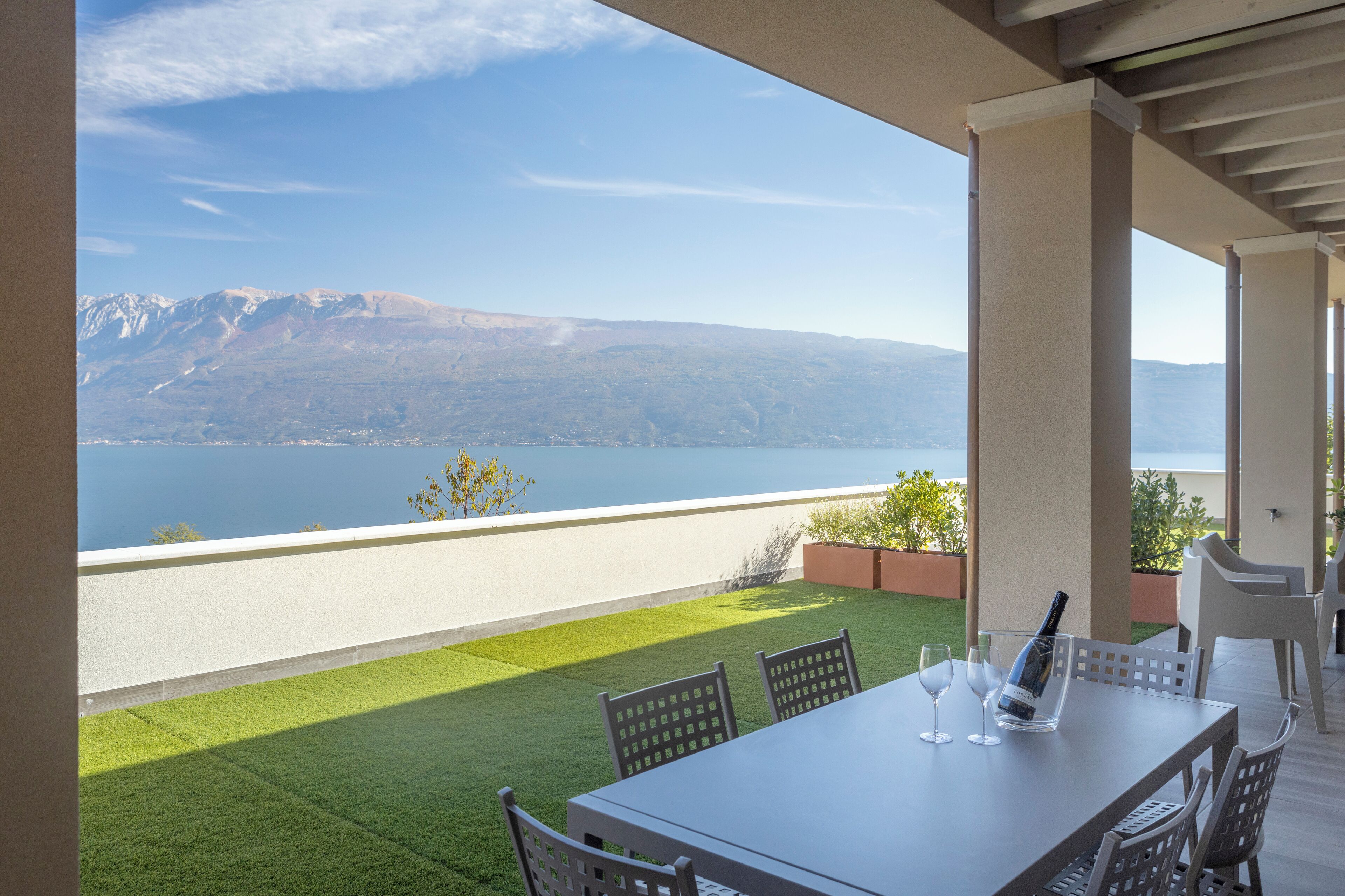 Une terrasse avec une table et des chaises, une bouteille de champagne et une vue sur un lac et des montagnes.