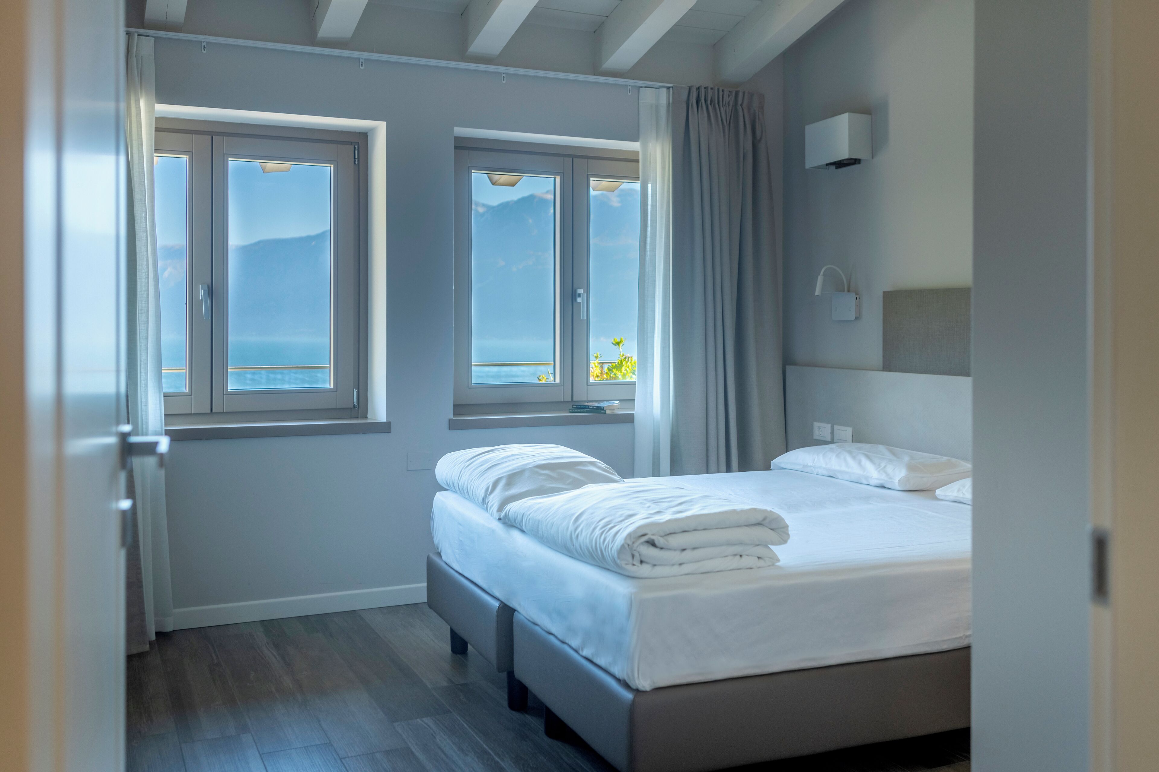 A modern hotel room with a bed, two windows, a view of the sea, and a ceiling fan.