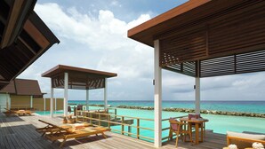 A wooden deck with lounge chairs and a table, overlooking a turquoise sea and a rocky shoreline. - Meyyafushi Maldives - Premium All Inclusive (Meyyafushi)