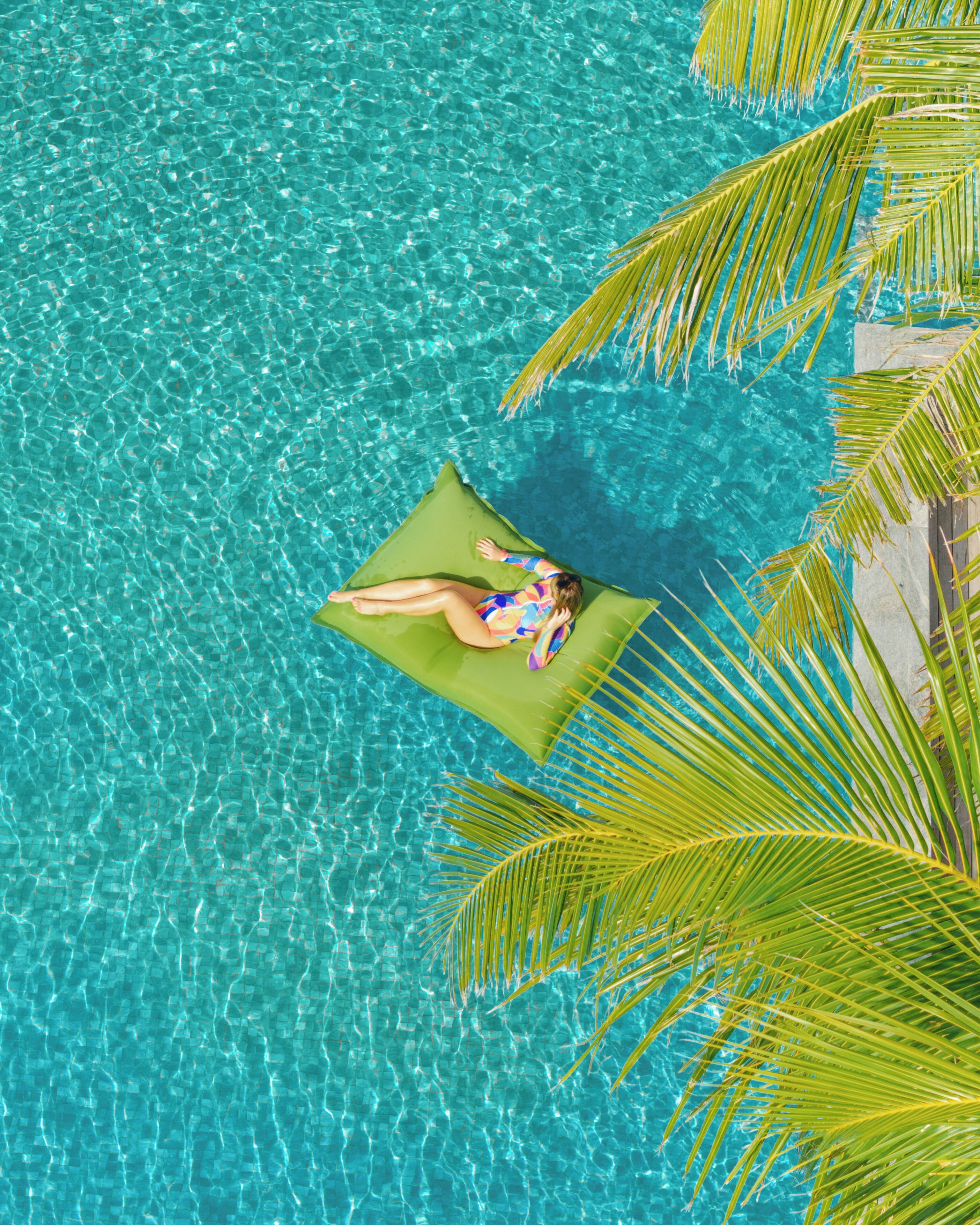 A person lying on a floating platform in a clear blue pool, surrounded by palm trees.