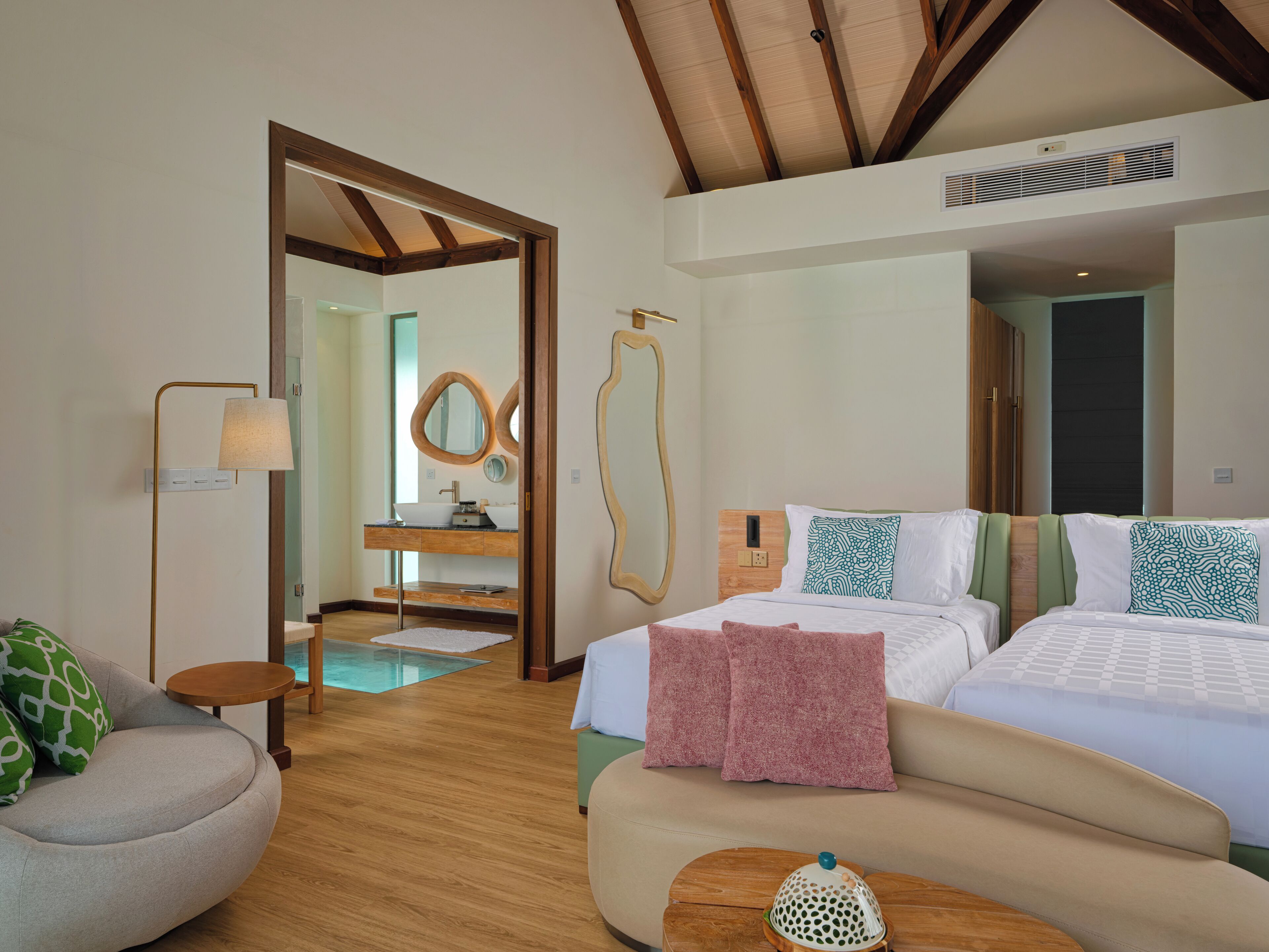 A hotel room with a wooden floor, a bed with white bedding, a sofa with green and pink pillows, a small round table, and a mirror with a wooden frame.