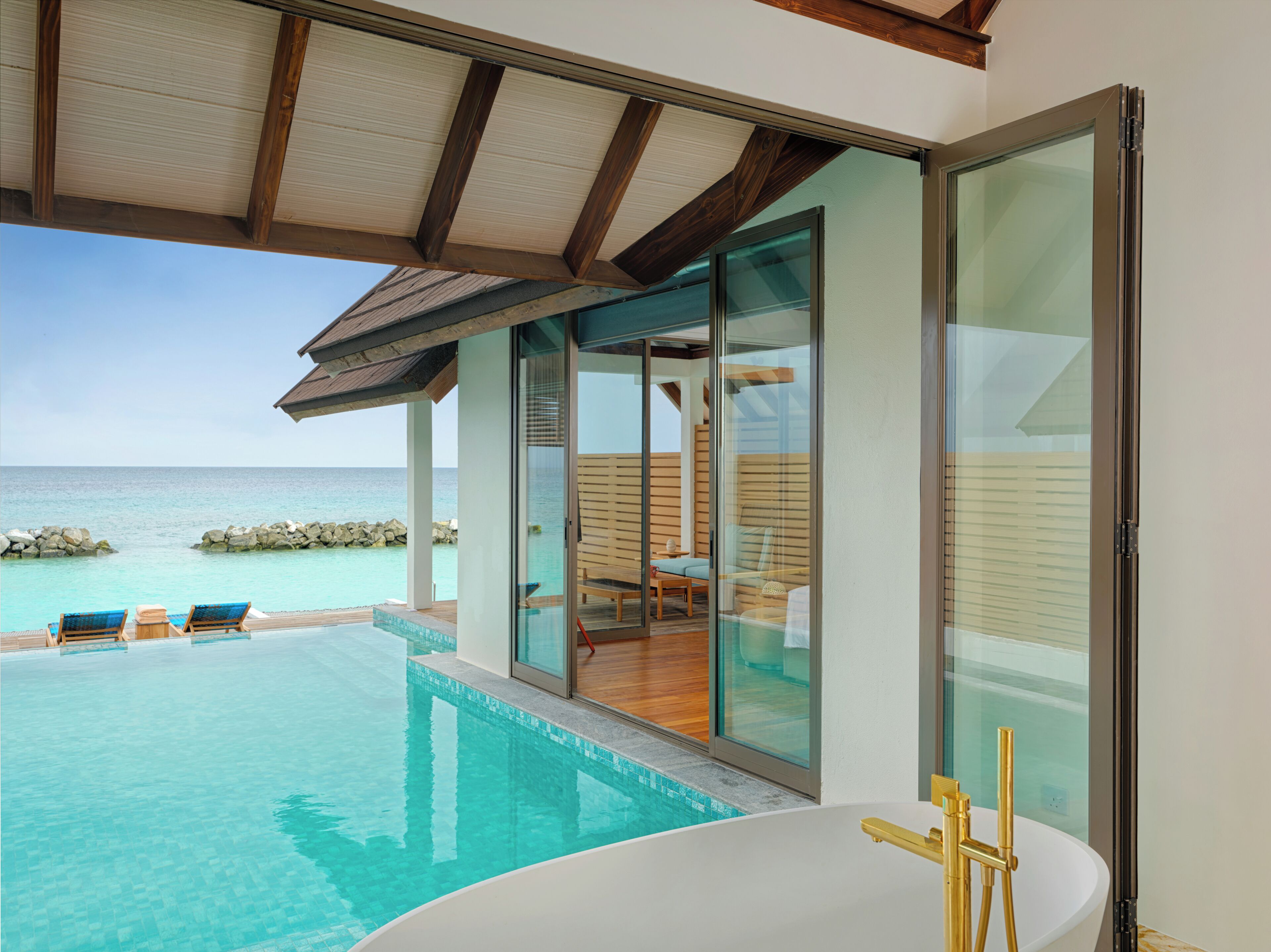 A modern infinity pool with a clear view of the ocean, a glass-enclosed seating area with a wooden deck, and a white infinity pool edge.