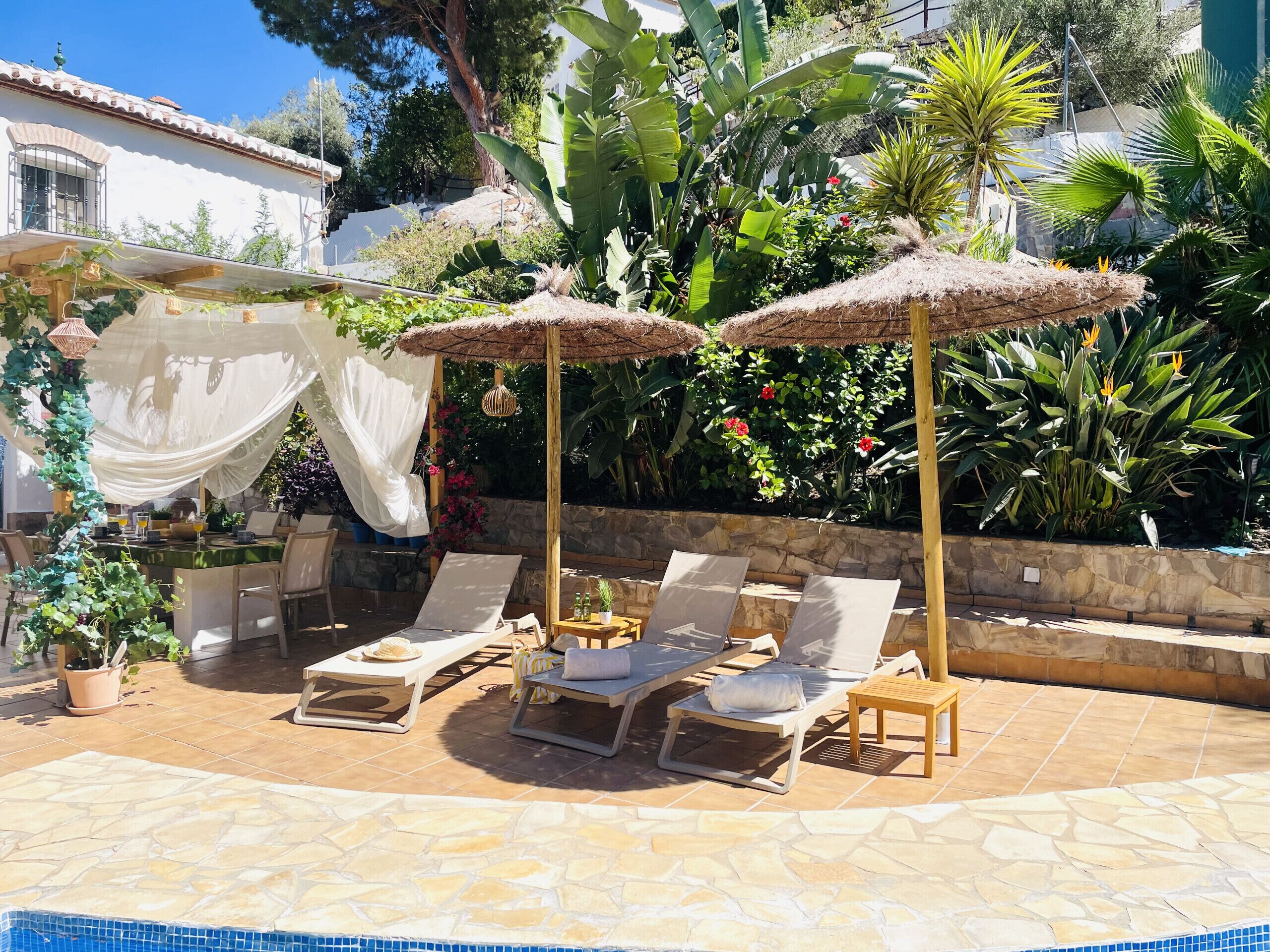 A pool area with lounge chairs and umbrellas, surrounded by lush greenery and a stone wall.