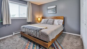 A bedroom with a bed, a window with curtains, a wall-mounted lamp, and a painting.