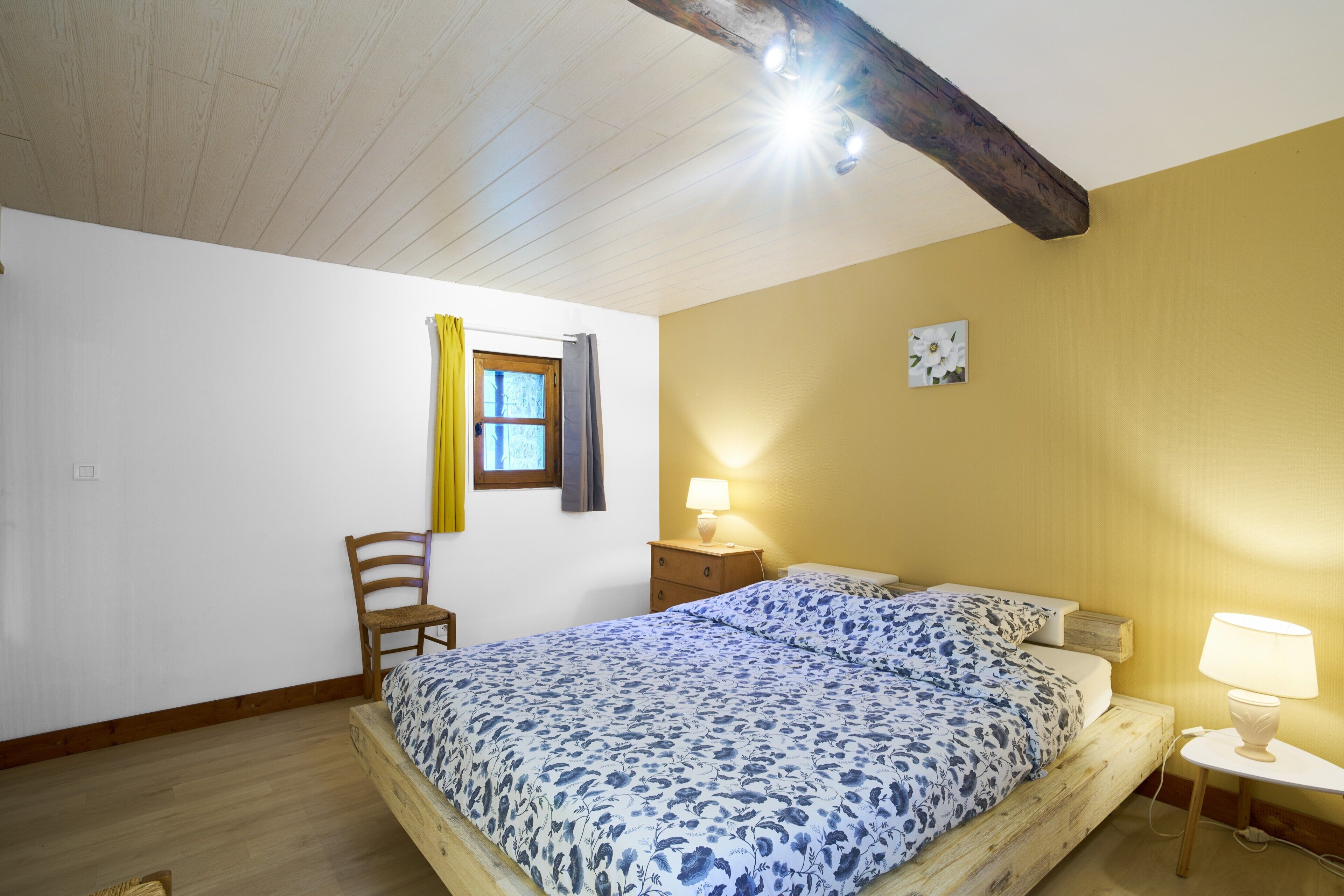 A bedroom with a wooden bed frame, a nightstand, a chair, a window with curtains, and a wall-mounted lamp.