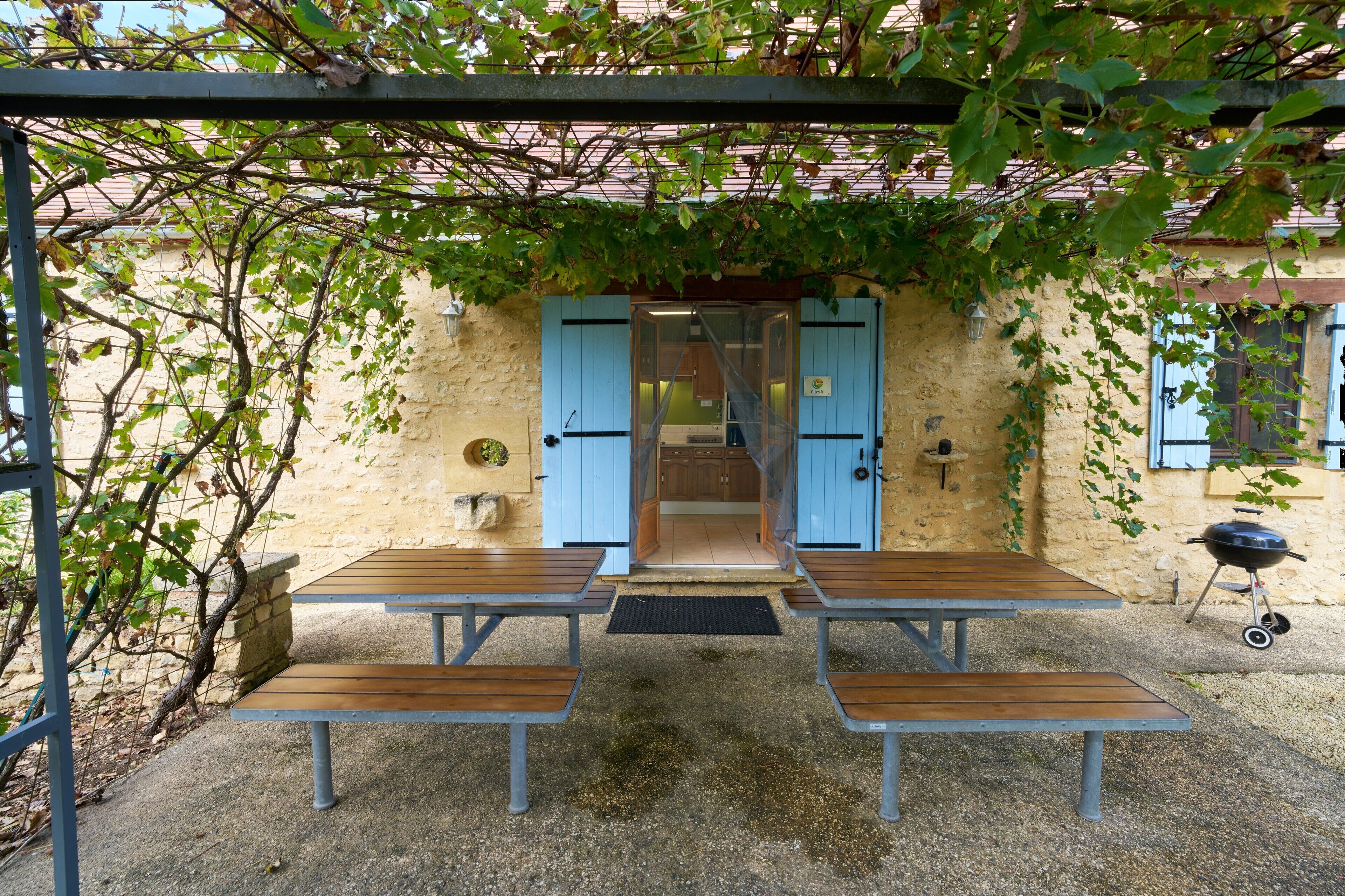 An outdoor seating area with tables and benches, a pergola, and a building entrance with blue doors.