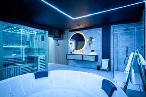 A modern bathroom with a large bathtub, a glass-enclosed shower area, and a vanity with a circular mirror. - Suite Ôna - Bassenge Wellness Suites (Bassenge)