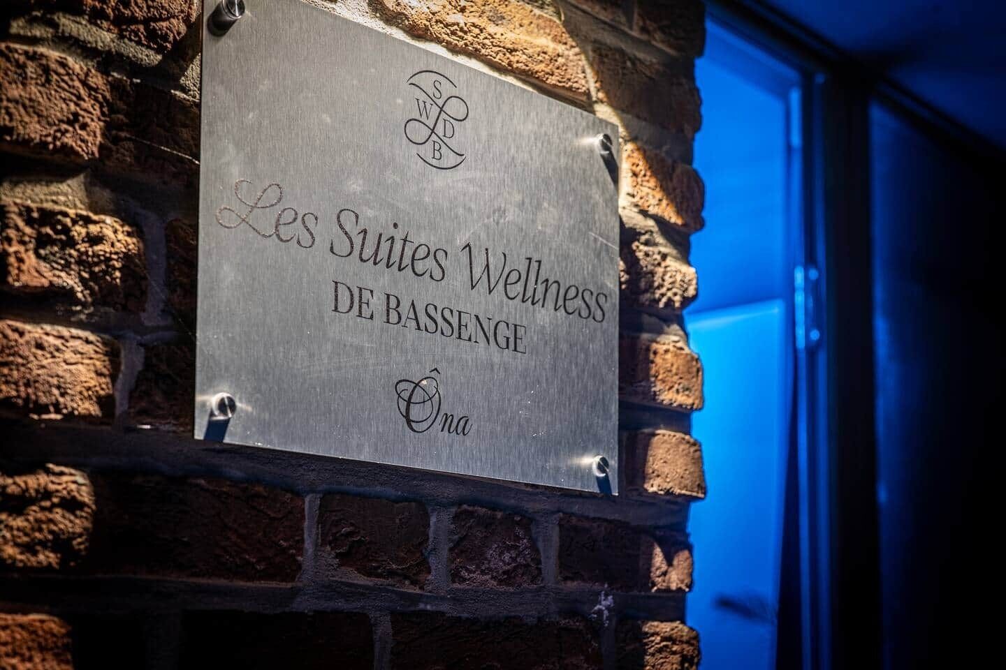 Une enseigne indiquant « Les Suites Wellness DE BASSENGE » est fixée à un mur de briques.