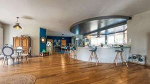 A spacious living area with a wooden floor, a dining table with chairs, a kitchen with a bar area, and a large window providing natural light. - 3 bedroom nice home in Sarliac (Sarliac)