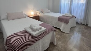 A hotel room with two beds, each with a striped bedspread and white pillows. There is a nightstand with a lamp between the beds, and a window with sheer curtains. - NEW! Ciudad de las Artes Apartment (València)