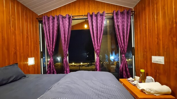 Ein Zimmer mit einem Bett, einem kleinen Tisch mit Pflegeprodukten und einem Fenster mit violetten Vorhängen.