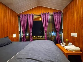 Quarto com cama, uma mesinha de apoio com artigos de higiene e uma janela com cortinas roxas.