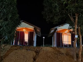 Duas pequenas cabanas de madeira com grandes janelas, localizadas numa trilha de terra com árvores de cada lado.