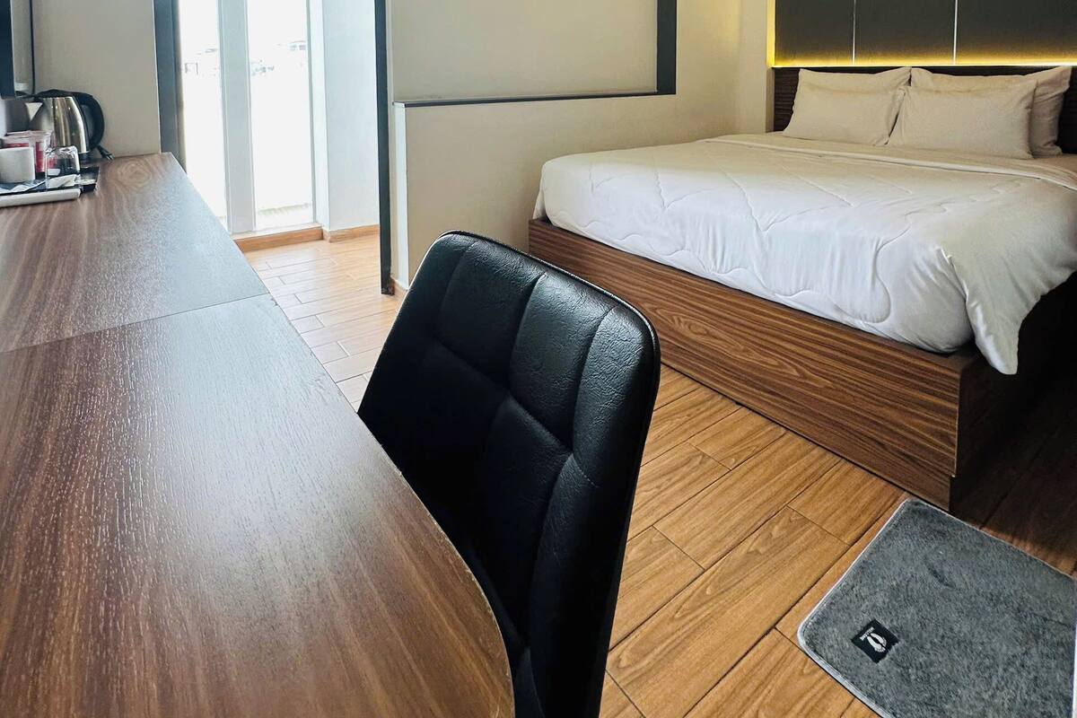 A hotel room with a wooden dining table, a single black chair, a bed with white bedding, and a large window.
