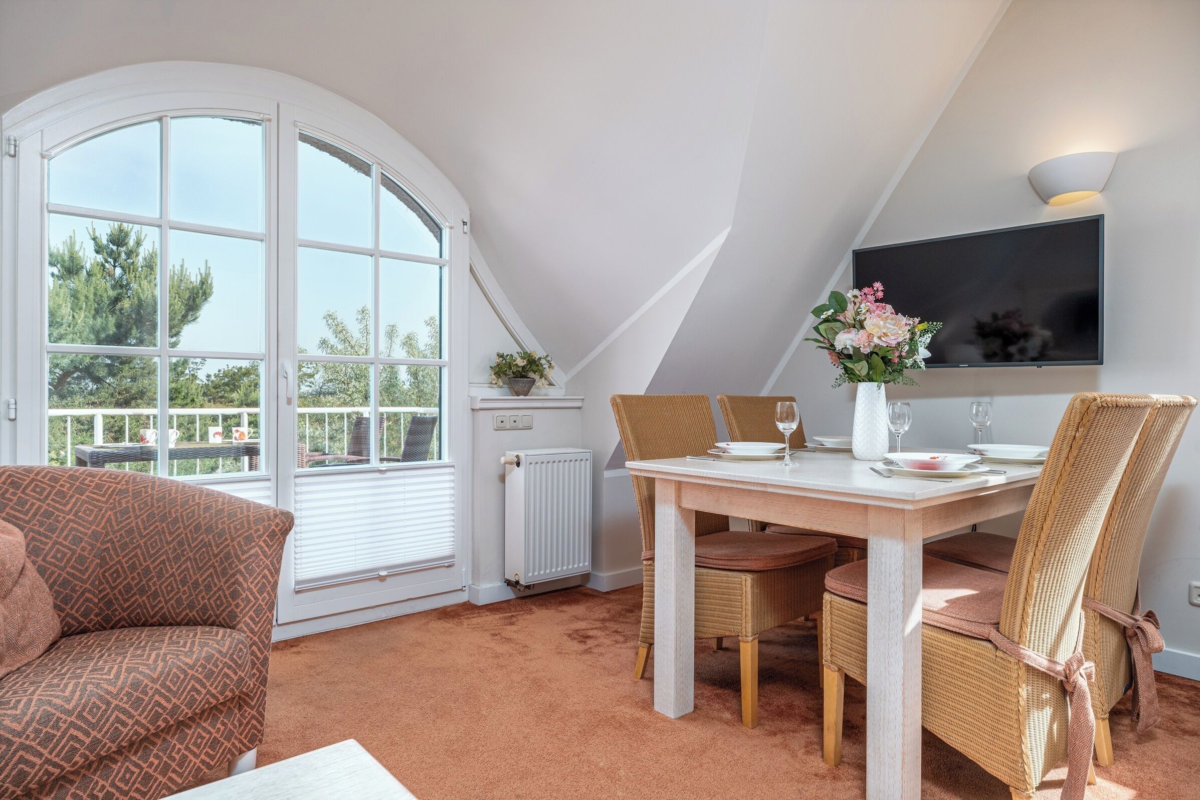 A cozy room with a large arched window, a dining table set for two, and a flat-screen TV mounted on the wall.