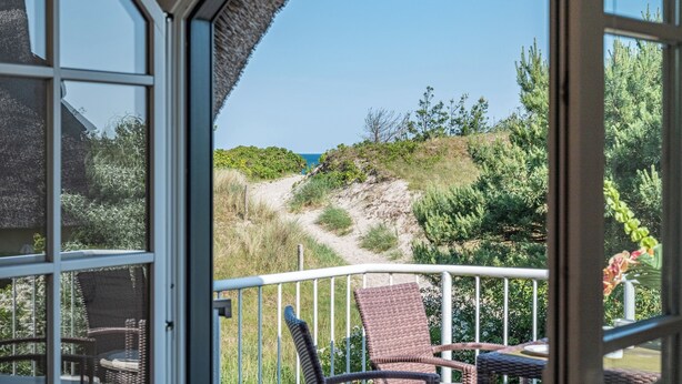 Un balcon avec des fauteuils en osier, une balustrade et une vue sur un sentier côtier bordé de verdure, sous un ciel dégagé.