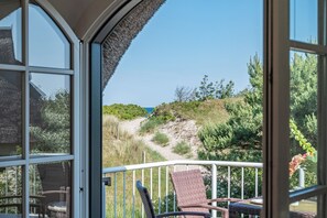 Un balcon avec des fauteuils en osier, une balustrade et une vue sur un sentier côtier bordé de verdure, sous un ciel dégagé.