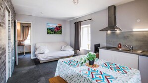 A compact living space with a bed, a dining table with a patterned tablecloth, a kitchen area with a stove and sink, and a painting on the wall.