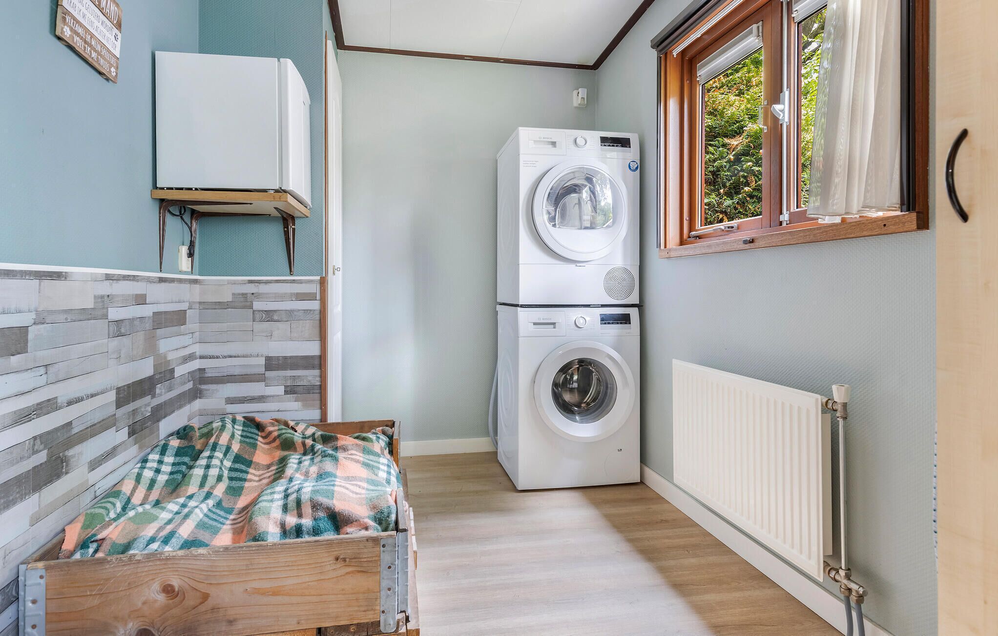 Une petite buanderie équipée d’un lave-linge et d’un sèche-linge, d’un radiateur et d’une fenêtre donnant sur un espace verdoyant.
