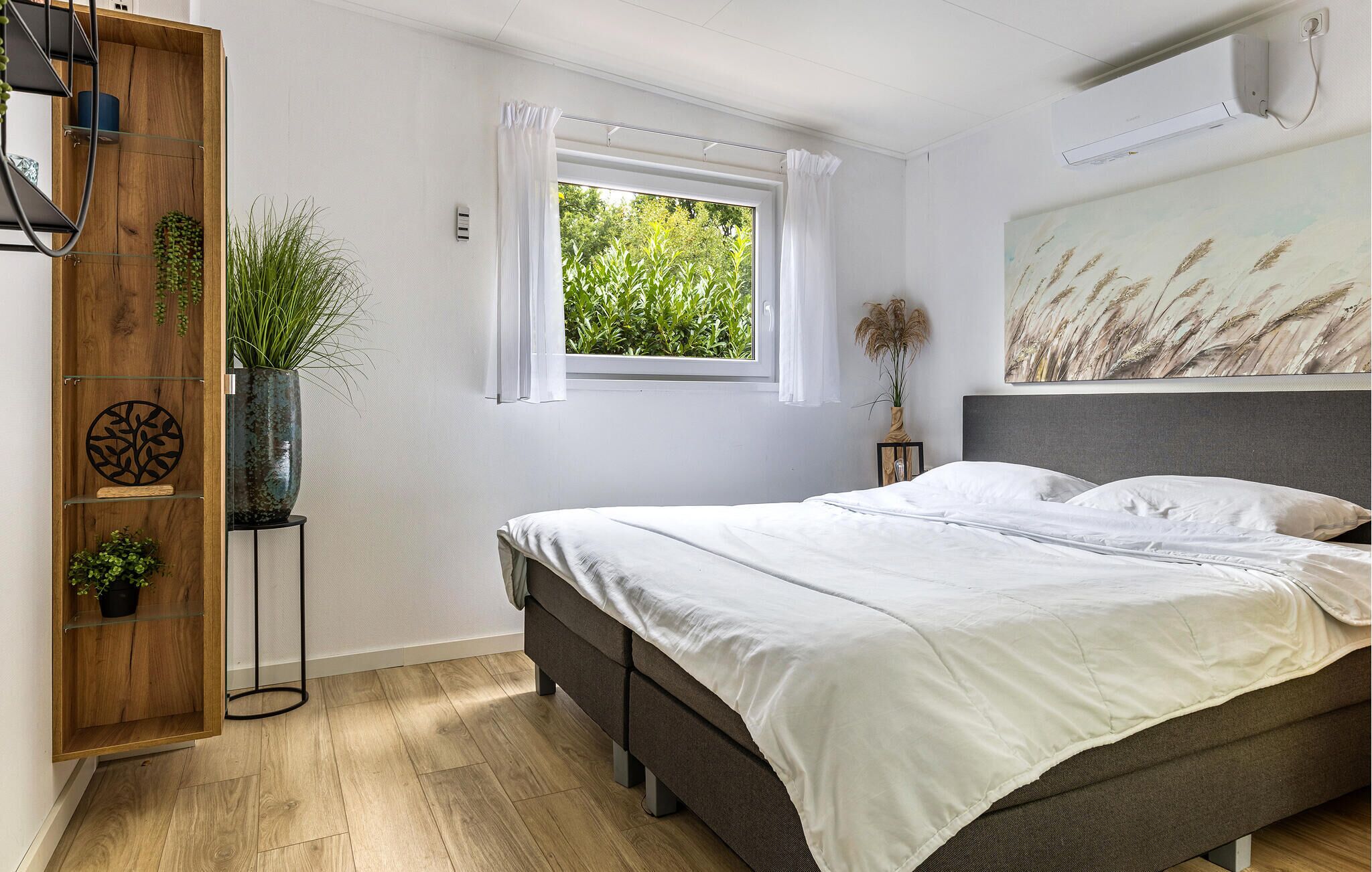 A bedroom with a bed, a window, a wooden wardrobe, and a wall-mounted air conditioner.