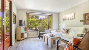 A modern living room with a sofa, a coffee table, a TV, and a desk with a lamp. There is a window with a view of greenery outside. - 2 bedroom pet friendly home in Erm (Erm)