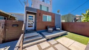Une maison moderne avec une porte d’entrée en bois, un petit porche agrémenté de mobilier d’extérieur et un ciel bleu dégagé.