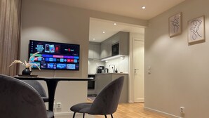 Un salon moderne avec un téléviseur à écran plat, des fauteuils gris et une petite table sur laquelle est posé un vase. On aperçoit une cuisine en arrière-plan, avec un four à micro-ondes et des placards.