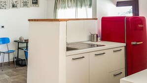 Una kitchenette compacta con un lavamanos, una hornilla pequeña y un refrigerador rojo. Se observa una silla azul y una mesa con un florero.