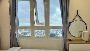 Ett hotellrum med en säng, ett fönster med gardiner, en rund spegel, ett litet bord med en vattenkokare och utsikt över en stad.