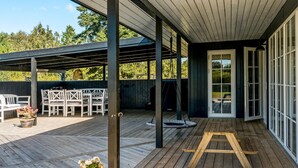 A covered outdoor deck with a wooden floor, white furniture, and a swing set.