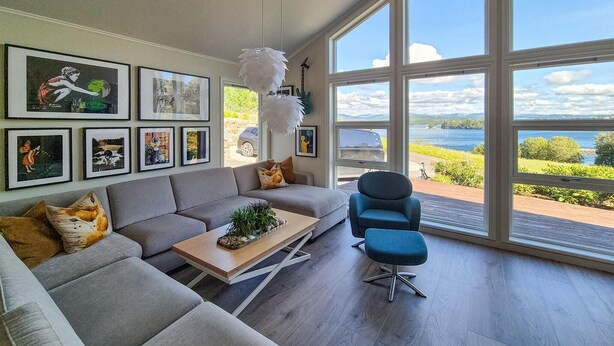 Una sala de estar moderna con un sofá gris, un sillón azul y una mesa de centro. Hay cuadros enmarcados en la pared y una vista de un lago a través de amplias ventanas.