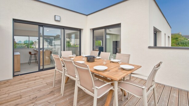 Un espace de restauration extérieur moderne, doté d’une table et de chaises en bois, de grandes portes-fenêtres coulissantes et offrant une vue sur le paysage urbain.