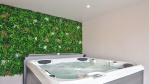 Une salle de spa moderne dotée d’un jacuzzi, d’un banc en bois et d’un mur recouvert de feuillage artificiel.