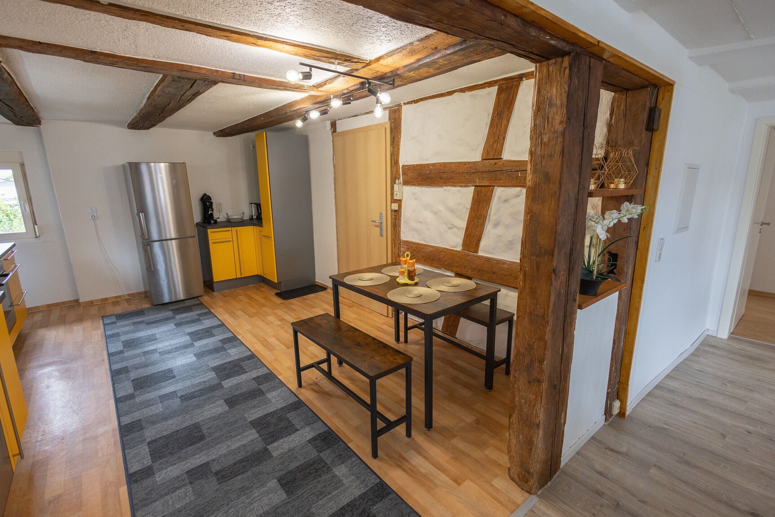 Une cuisine avec des poutres en bois, un réfrigérateur en acier inoxydable, des meubles de couleur jaune, une table à manger avec des sets de table et un banc.