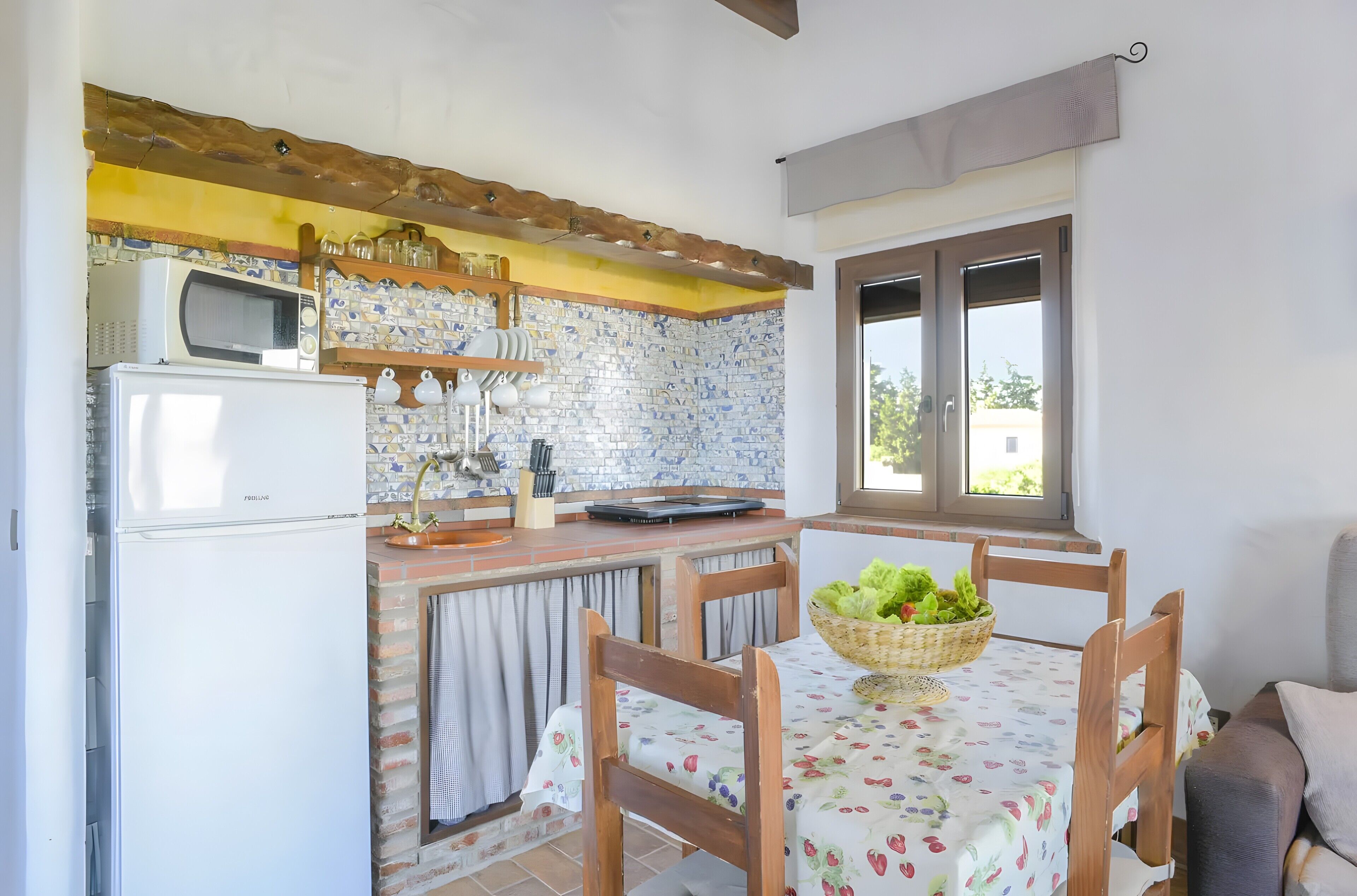 Una cocina con microondas, refrigerador y un salpicadero de ladrillo. Hay una mesa de comedor con sillas y una cesta de fruta.