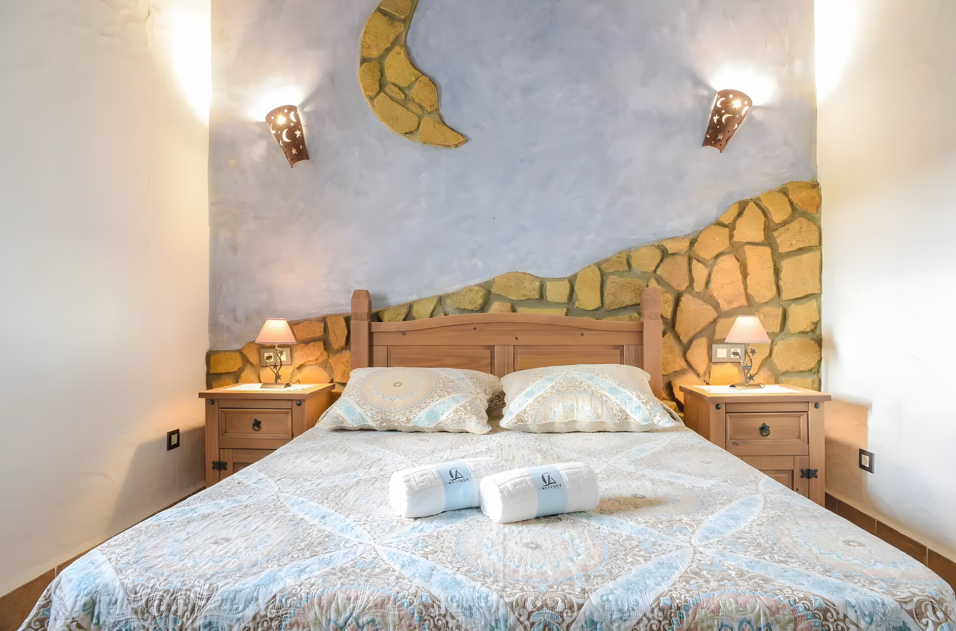 Una cama con una colcha estampada, dos lámparas de noche y una pared con diseño de luna creciente.