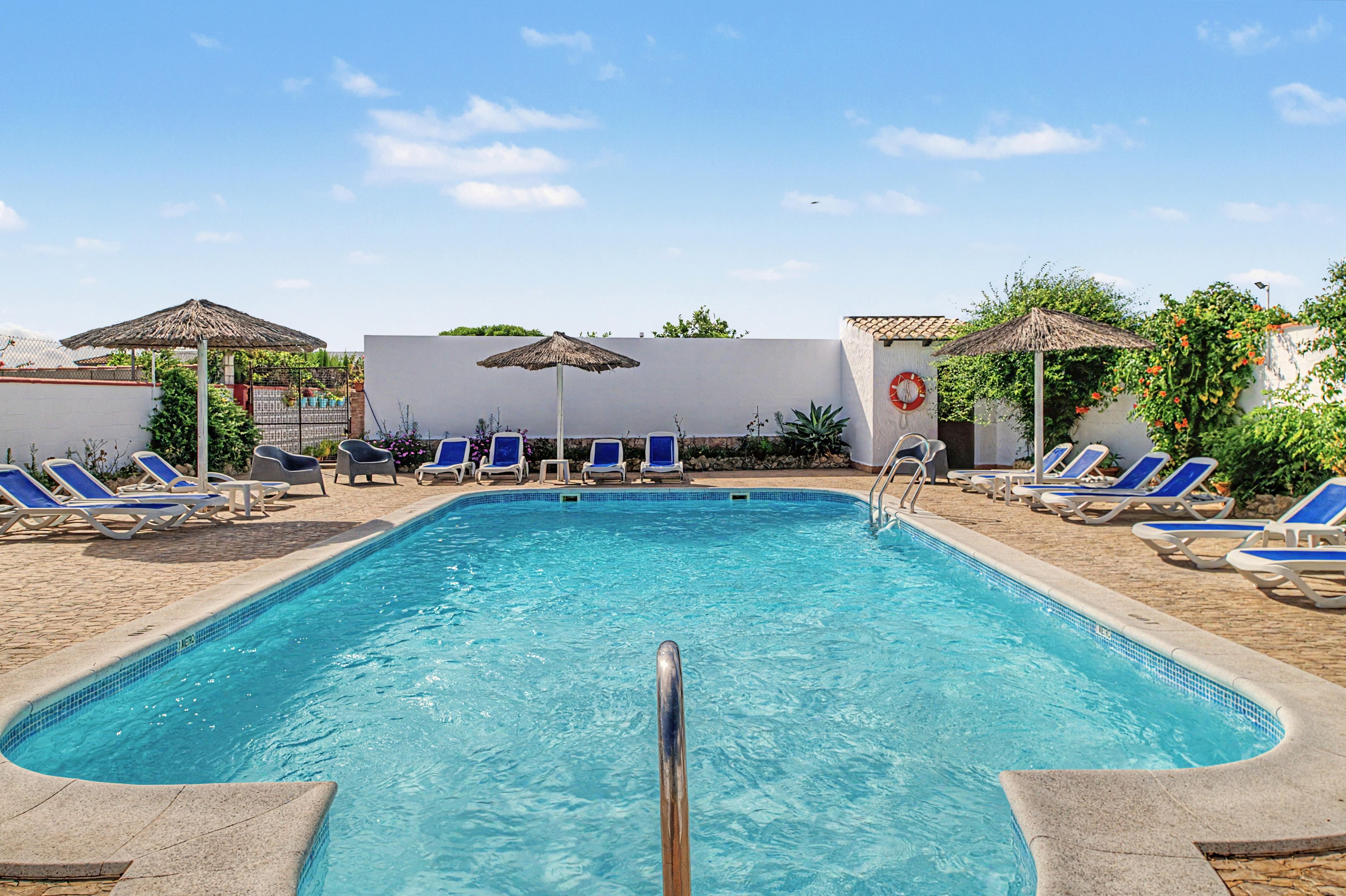 Ein Swimmingpool mit blauen Liegestühlen und Sonnenschirmen, umgeben von einer weißen Mauer und viel Grün.