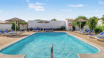 Ein Swimmingpool mit blauen Liegestühlen und Sonnenschirmen, umgeben von einer weißen Mauer und viel Grün.