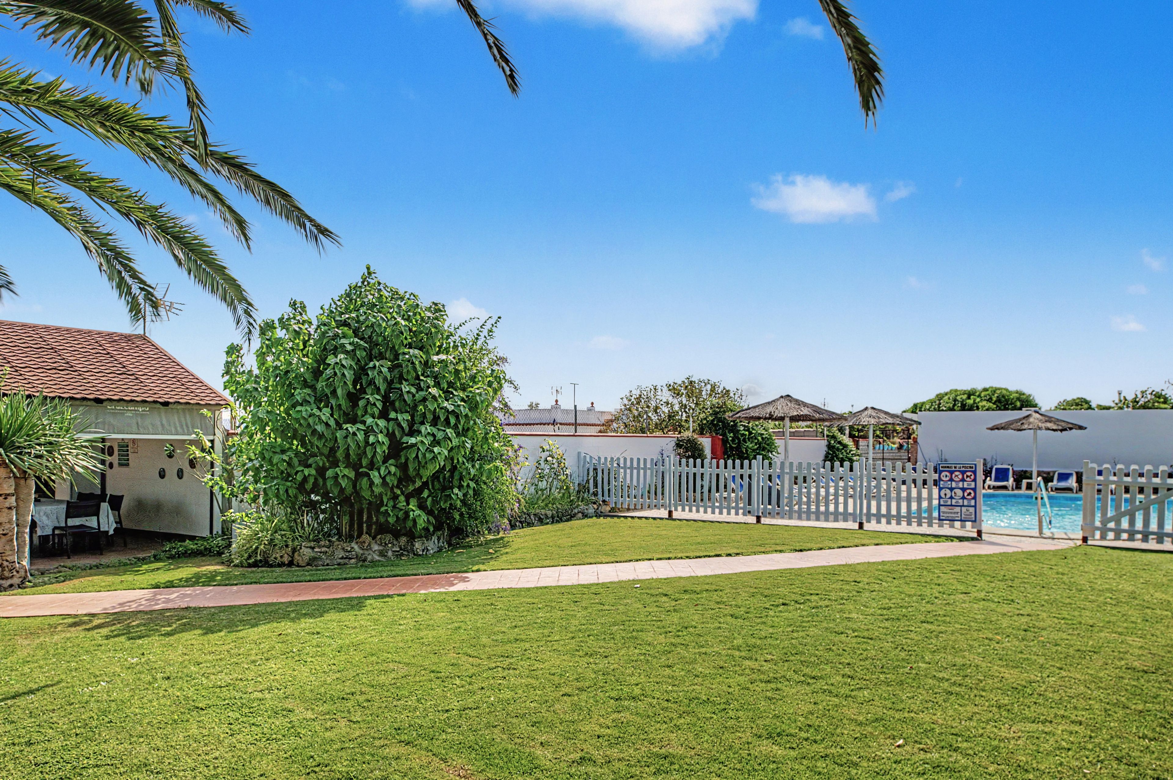 A pool area with a white fence, a grassy lawn, and a building with a tiled roof.