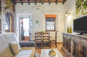 Un salon comprenant un canapé, un téléviseur posé sur un meuble en bois, un coin repas avec des chaises et une table, et un coin cuisine avec une cuisinière.
