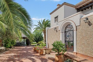 A courtyard with a paved walkway, palm trees, and a building with a stone facade and a metal gate. - Apartment 'El Nogal' with Shared Pool, Wi-Fi and Air Conditioning (Barbate)