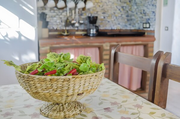 Un saladier en osier posé sur une table recouverte d’une nappe à motifs floraux, une chaise en bois et, au fond, une cuisine.