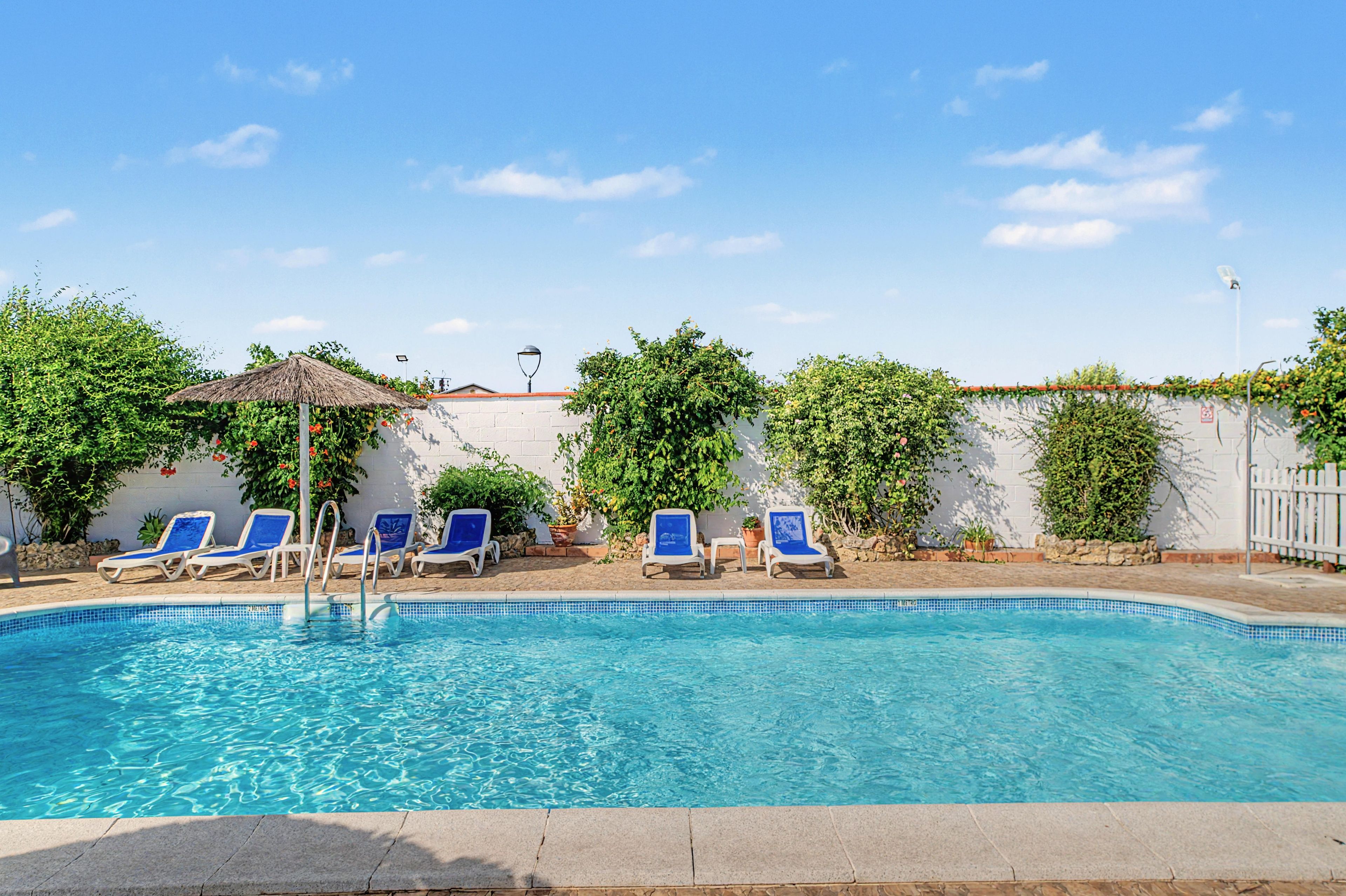 A swimming pool with clear blue water, surrounded by white walls and a white fence. There are several blue lounge chairs aligned along the poolside.