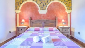 A bedroom with a stone wall, a wooden headboard, two bedside tables with lamps, a bed with a patterned bedspread, and two rolled towels on the bed. - Apartment 'Castaño' with Shared Pool, Wi-Fi and Air Conditioning (Barbate)
