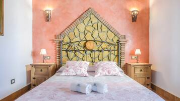 Quarto com cabeceira estampada em pedra, duas mesas de cabeceira com luminárias, cama com roupa de cama floral rosa e duas toalhas enroladas na cama.